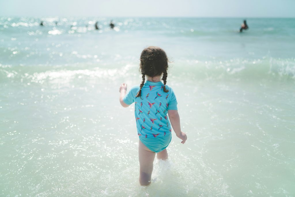 Girl with dark hair, walking into the sea on a sunny day, wearing a blue patterned sun safe swimsuit